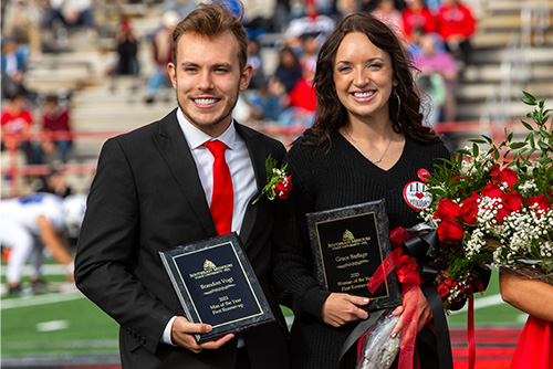 A man and woman stand and smile together, holding flowers and awards