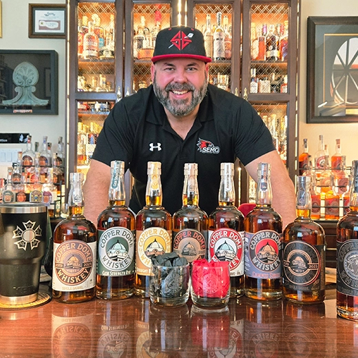 A man in a polo and baseball cap stands at a wooden table with Copper Dome Whiskey offerings. 
