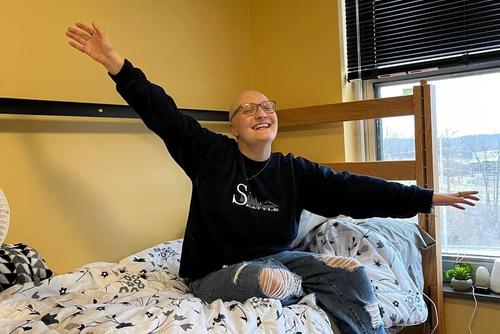 Gianna Deidrick sits on her dorm room bed smilling with arms wide.