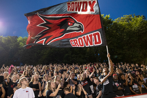 A crowd of students cheer at a football game while waving a Rowdy Crowd flag.