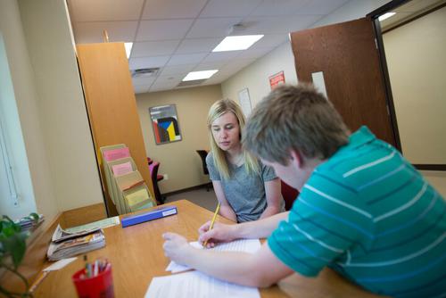 Two people seated at a desk in front of a window, both looking at a typed paper on the table, one is making editing marks