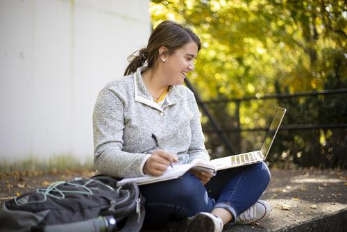 A student takes notes and looks at a computer while studying outdoors on campus.