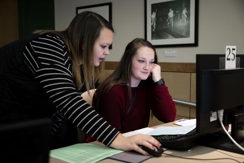 A staff member advises a student as they work at a computer.