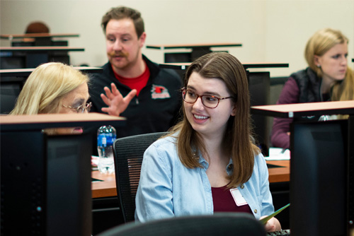 A student smiles while sitting at a computer and receiving advisement from a staff member.