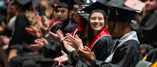 A line of students seated at graduation are clapping and smiling during the commencement ceremony.