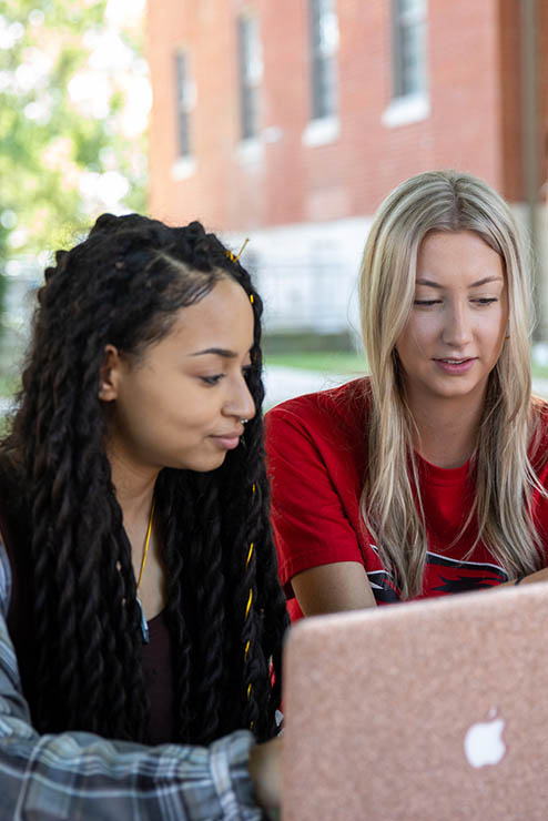 two students sitting together outside at a picnic table under a shade tree studying together
