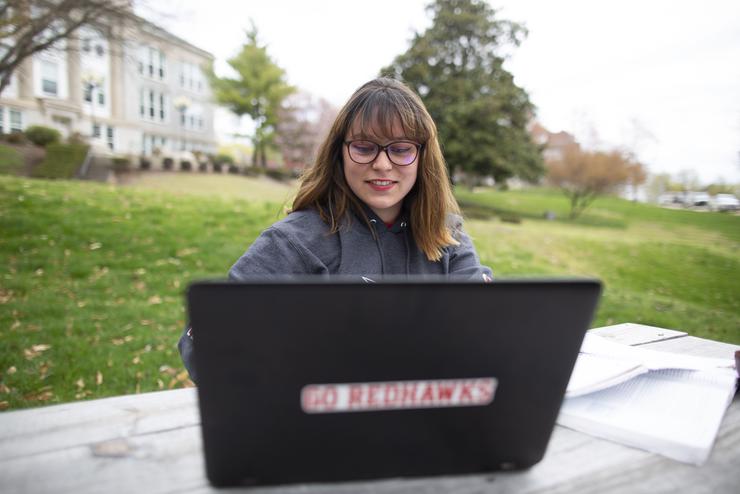 A student sits at a picnic table and works on their laptop that has a redhawks sticker on the back. 