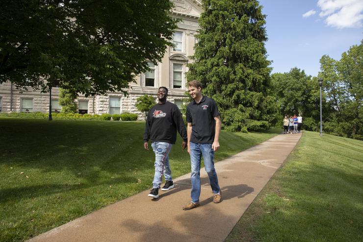 Two students walk on the sidewalk in front of Academic Hall. 