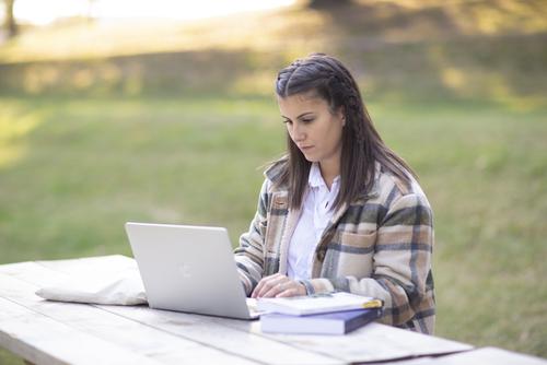 A student sits at a wood picnic table in a grass area with a laptop and two textbooks.