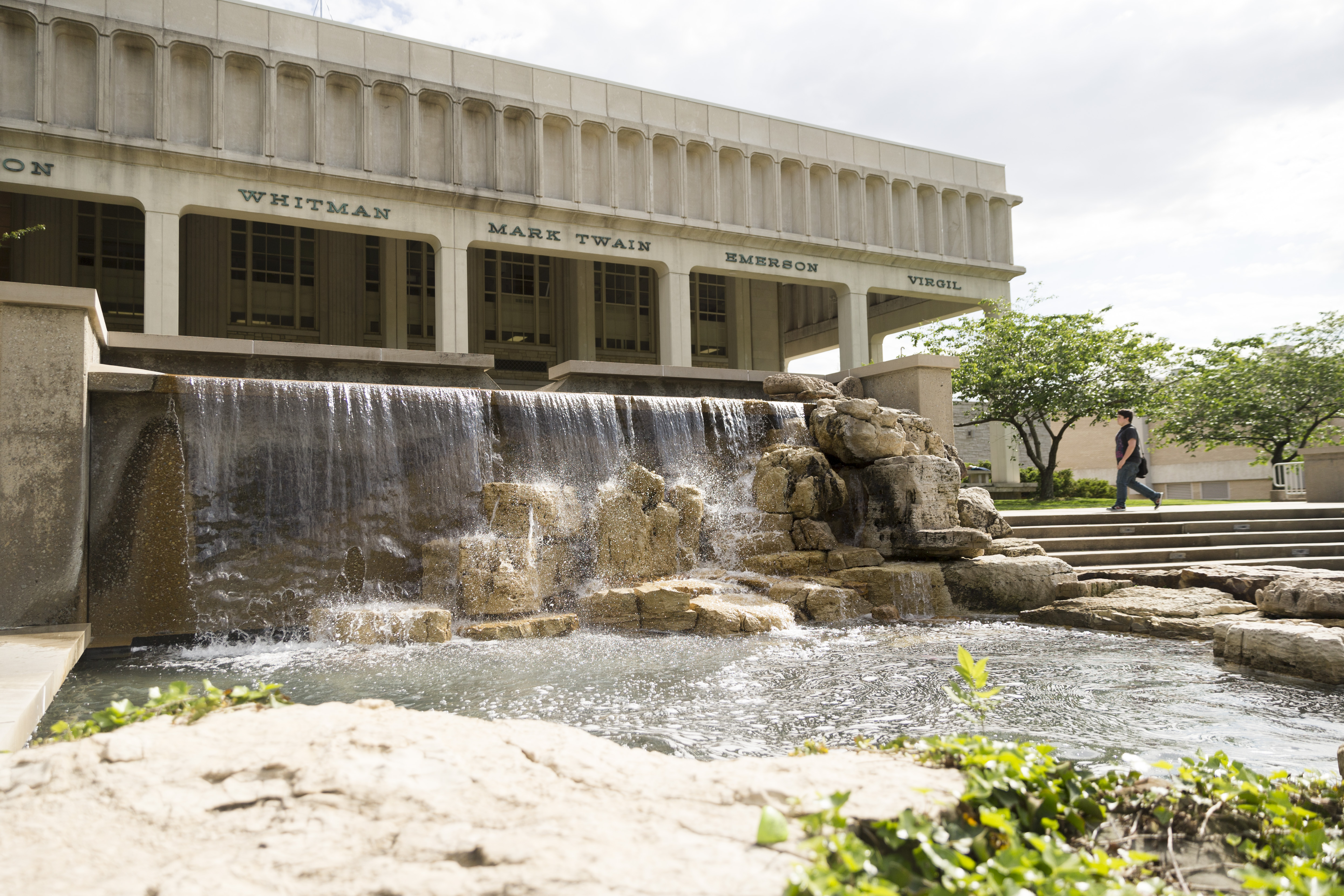 the rocky waterfall fountain in front of the Kent Library veranda, a student crosses the sidewalk in the background